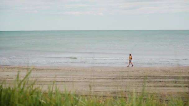 Mulher de casaco amarelo e calções jeans caminha ao longo da costa do Mar Mediterrâneo. Plano geral — Vídeo de Stock