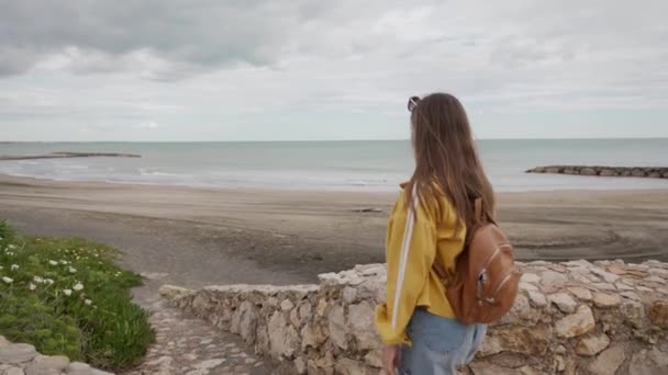 在恶劣的天气里，背着背包的女游客走在一条通向美丽蓝色大海的石路上。 后视镜 — 图库视频影像