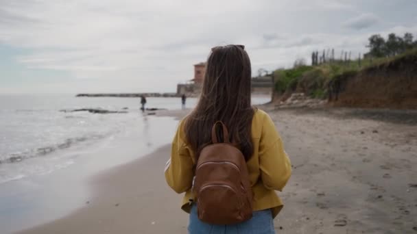 Chica turística con mochila marrón caminando a lo largo de la playa del Mar Mediterráneo en el día de primavera. Vista trasera — Vídeo de stock