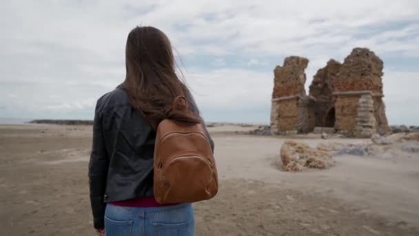 Chica morena de pelo largo con una mochila se acerca a las ruinas de una antigua fortaleza Torre Flavia en Ladispoli, Italia. Vista trasera — Vídeos de Stock