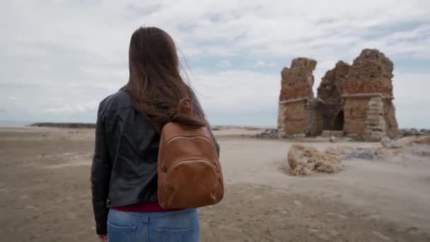 Fille brune aux cheveux longs avec un sac à dos s'approche des ruines d'une ancienne forteresse Torre Flavia à Ladispoli, Italie. Vue arrière — Video