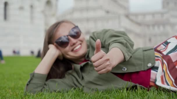 Portret młodej kobiety w okularach przeciwsłonecznych relaksującej się na trawniku baptysterium św. Jana na Placu Cudów i pokazującej coś w tym stylu. Piza, Włochy — Wideo stockowe
