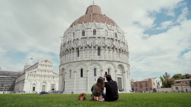 Joven hombre y mujer sentado y tomando fotos del famoso edificio de Pisa utilizando el teléfono móvil. Vista trasera. Baptisterio de San Juan. Vacaciones en Italia, temporada de viajes — Vídeo de stock