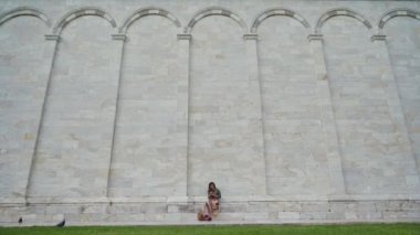 İtalya, Piazza dei Miracoli 'de ünlü Campo Santo' nun mermer duvarının yanında oturan güzel kız. Genel görünüm