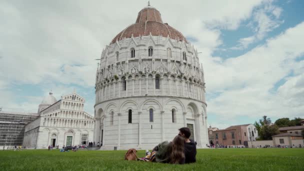 Pareja de turistas que se encuentran en la hierba frente al Baptisterio de San Juan y mirando a la famosa marca en Pisa. — Vídeos de Stock