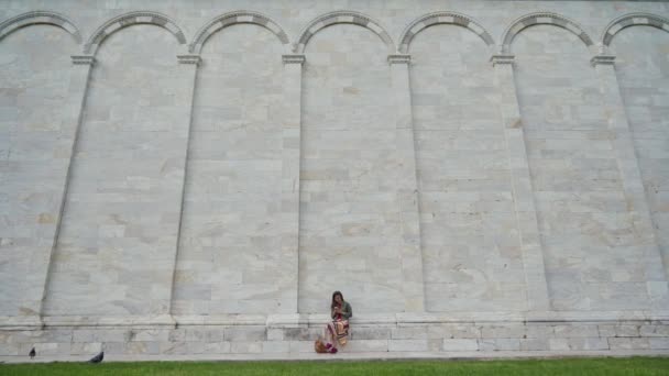 Piękna dziewczyna siedzi za pomocą telefonu komórkowego w pobliżu marmurowej ściany słynnego zabytku Campo Santo na Piazza dei Miracoli, Włochy. Ogólny pogląd — Wideo stockowe