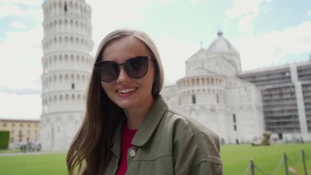 Junge hübsche Frau mit Sonnenbrille steht auf der Piazza dei miracoli vor der Kathedrale von Pisa und dem schiefen Turm von Pisa und lächelt an einem schönen Tag in die Kamera — Stockvideo