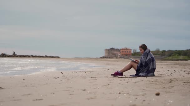 Romantic girl reads book while sitting by sea on cool spring day. Side view — 图库视频影像