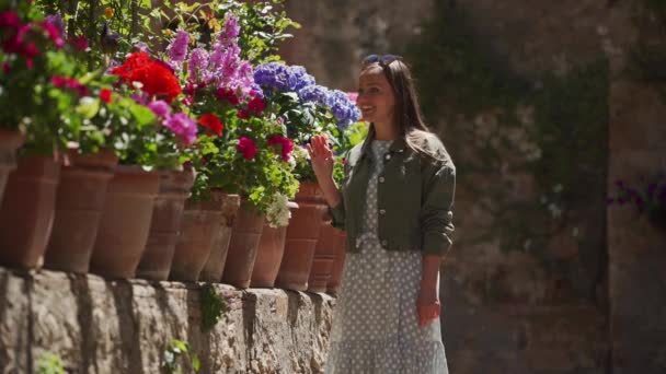 Happy girl in a romantic dress, jacket walks along flower bed and touches flower — Stockvideo