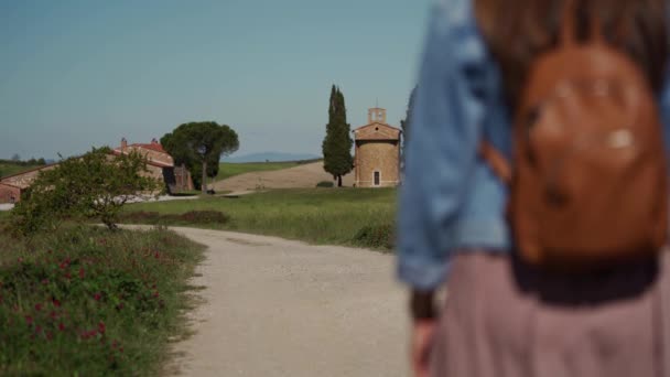 Woman in dress, denim jacket walking along path to chapel. Long shot. Rear view — Stockvideo