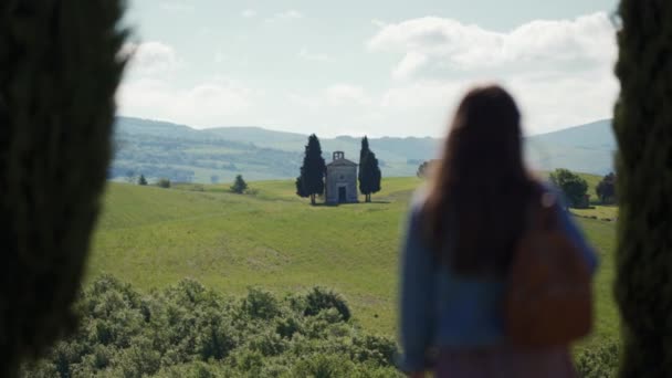 Girl with backpack stands, enjoys scenic Tuscan landscape with Vitaleta chapel — 图库视频影像