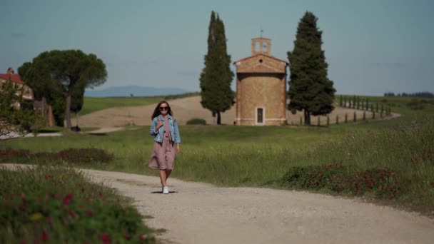 Young girl in sunglasses, dress and jacket walks along path on chapel background — Stockvideo