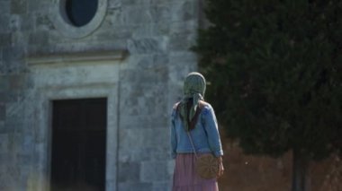 Beautiful girl in a scarf looks at small chapel, turns to camera, sweetly smiles