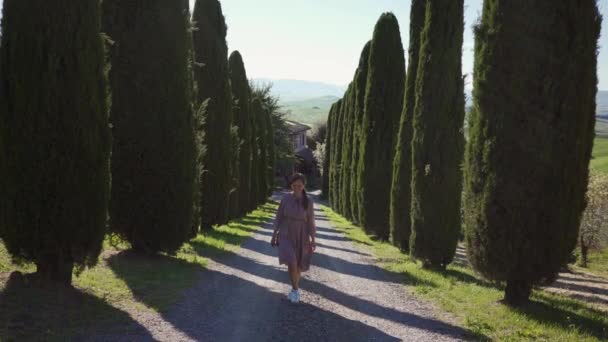 Happy girl in romantic beige dress walks along cypress alley on sunny day. Front — ストック動画