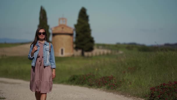 Menina em óculos de sol, vestido e jaqueta caminha ao longo do caminho no fundo da capela borrão — Vídeo de Stock