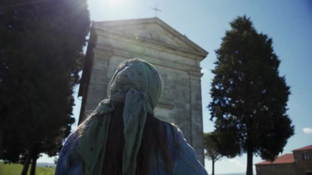 Woman in scarf stands near Cappella di Vitaleta, cross herself. Sun rays, chapel — ストック動画