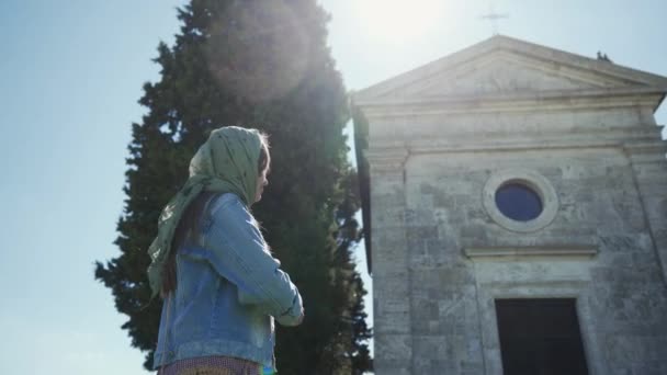 Woman in scarf stands near Cappella di Vitaleta, cross herself. Side view, sunny — ストック動画