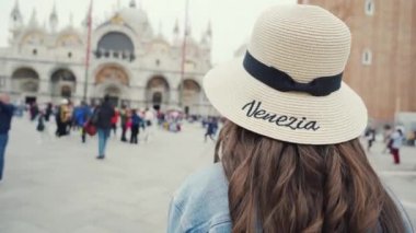 Hasır şapkalı güzel kız San Marco meydanında yürüyor, arkasına bakıyor ve gülümsüyor.