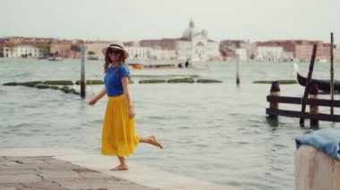 Şapkalı mutlu bir kız, sarı etekli yalınayak koşuyor, salınıyor, Venedik 'te iskelede eğleniyor.
