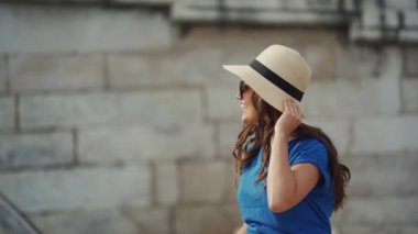 Şapkalı kız suyun yanında yalınayak dinleniyor. Gülümsüyor, elinde ayakkabılarla dans ediyor.