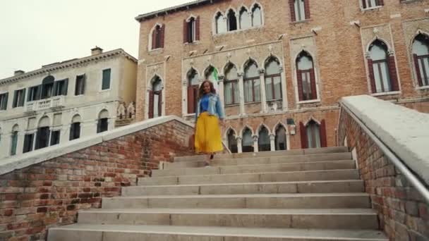 Брюнетка в желтой юбке спускается по лестнице по мосту через канал в Венеции — стоковое видео
