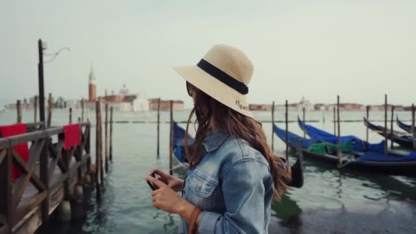Meisje toerist in zonnebril, hoed neemt foto per telefoon van gondels op Grand Canal — Stockvideo