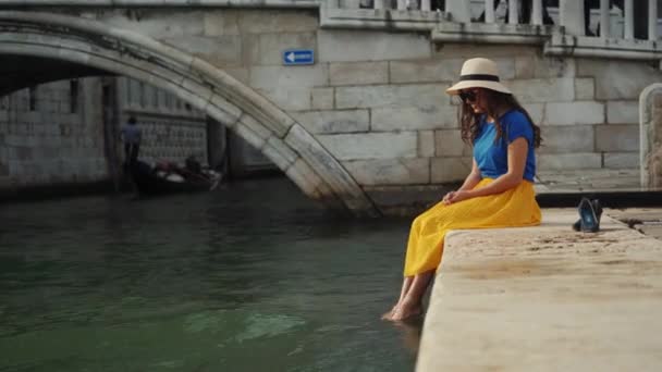 Gelukkig vrouw in hoed zit op pier en maakt spatten water met haar blote voeten — Stockvideo