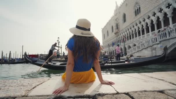 Venedig, Italien - 21 maj 2019: Brunett flicka i hatt sitter på piren, ser gondoler — Stockvideo