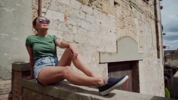 Woman in sunglasses, denim shorts sits on stone fence in old Italian city, sunny — Stock Video