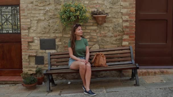 Bonito jovem turista menina sentar no banco de madeira perto de parede de tijolo velho com vasos de flores — Vídeo de Stock