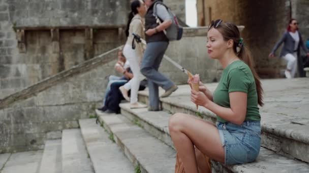 Vrouw met bril eet Italiaans ijs met aardbei in kegel zittende stappen — Stockvideo