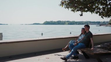 Birbirine aşık bir çift, gezinti alanındaki ağacın altında oturmuş Garda Gölü manzarasına hayran.