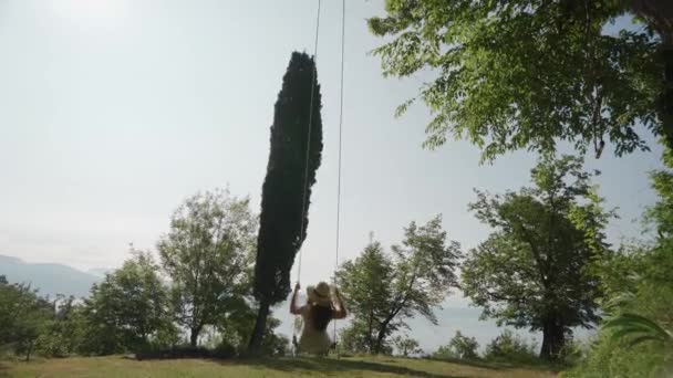 Mujer en vestido, sombrero de paja se balancea en columpio con vista al lago, vista trasera, soleado — Vídeos de Stock