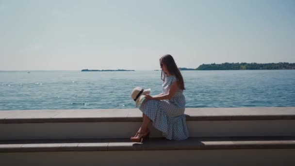 Donna in abito romantico siede vicino lago blu sul parapetto di pietra, tiene il cappello in mano — Video Stock