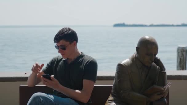 GARDONE RIVIERA, ITALY - MAY 23, 2019: man reads phone near statue, mirror pose — Stock Footage GARDONE RIVIERA, ITALY - MAY 23, 2019: man reads phone near statue, mirror pose Stock Video