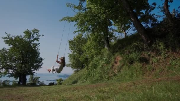 Leuke vrouw in zomerjurk, strohoed schommels op boom schommel, zijaanzicht. Grote kans. — Stockvideo