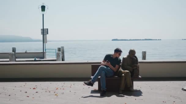 GARDONE RIVIERA, ITALY - MAY 23, 2019: guy sits on bench near statue, poses like — Stock Video