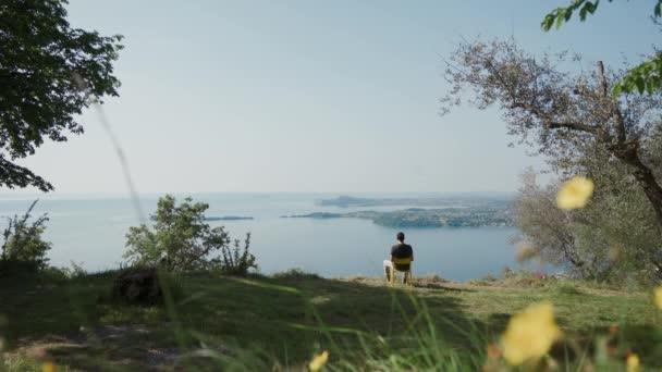 El joven se sienta en la silla amarilla en la colina, descansa, mira el hermoso paisaje del lago — Vídeos de Stock
