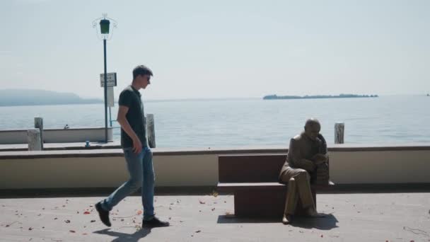 GARDONE RIVIERA, ITALY - 23 мая 2019 года: мужчина идет на скамейку со статуей, садится — стоковое видео