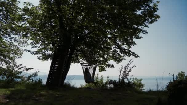 Silueta de niña descansando a la sombra del árbol en hamaca sobre el lago de Garda. Largo plazo — Vídeos de Stock