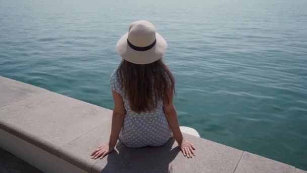 Vista posteriore della donna in cappello si siede e si rilassa sul molo di pietra sul lago nella giornata di sole — Video Stock