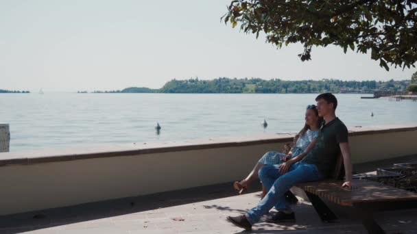 Ζευγάρι ερωτευμένο κάθεται στον πάγκο κάτω από το δέντρο σε περιπάτους, θαυμάζει τη θέα της λίμνης Garda — Αρχείο Βίντεο