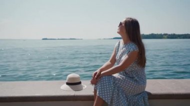 Güneş gözlüklü kadın şapkasının yanında oturuyor, Garda Gölü 'nün yanındaki taş iskelede dinleniyor.