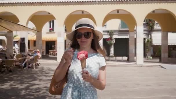 Girl in hat and sunglasses walk and eats red ice cream cone on arcade background — Stock Video