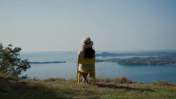 Mujer joven en sombrero se sienta en la silla en la colina, descansa, mira el paisaje del lago escénico — Vídeos de Stock