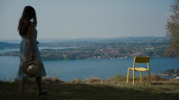 Chica en vestido con sombrero en la mano ir a la silla, se sienta a disfrutar de la vista del lago azul — Vídeos de Stock