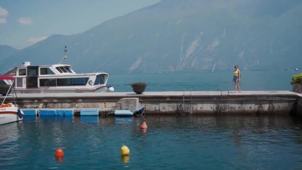 Chica turística camina sobre muelle de piedra lago con barco, contra de las montañas de los Alpes. Largo — Vídeo de stock