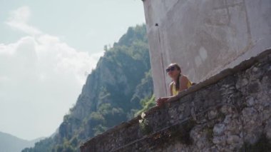 Güneş gözlüklü turist kadın taş terasta duran küçük kasaba manzarasından hoşlanıyor.