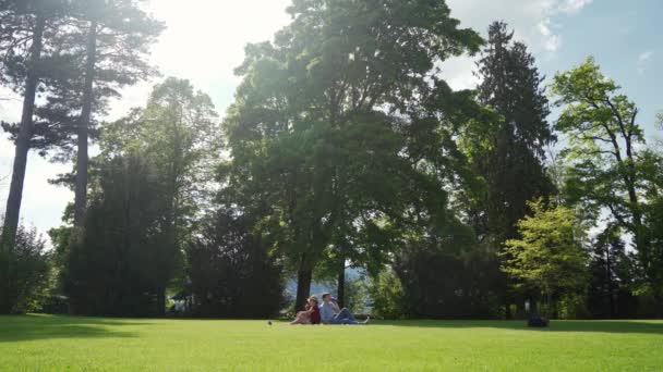 Ung familj sitter på gräs under picknick i vårparken under stora gröna träd — Stockvideo