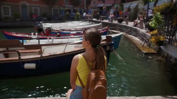Γυναίκα σε κίτρινο T-shirt με σακίδιο κάθεται στην προβλήτα με βάρκες, πίσω όψη, ηλιόλουστη — Αρχείο Βίντεο